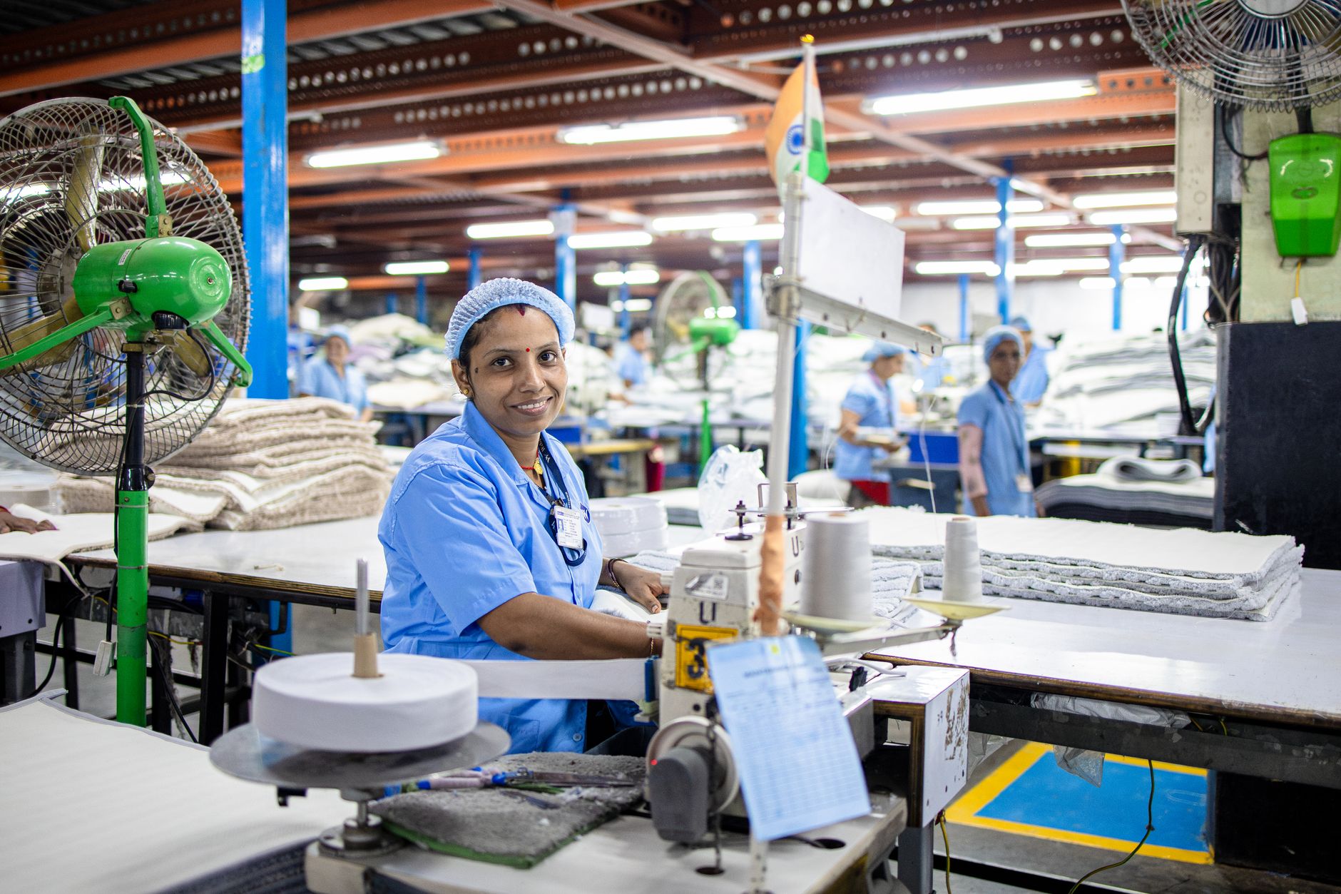 A female factory worker in India manages a sewing machine in a bustling textile factory.