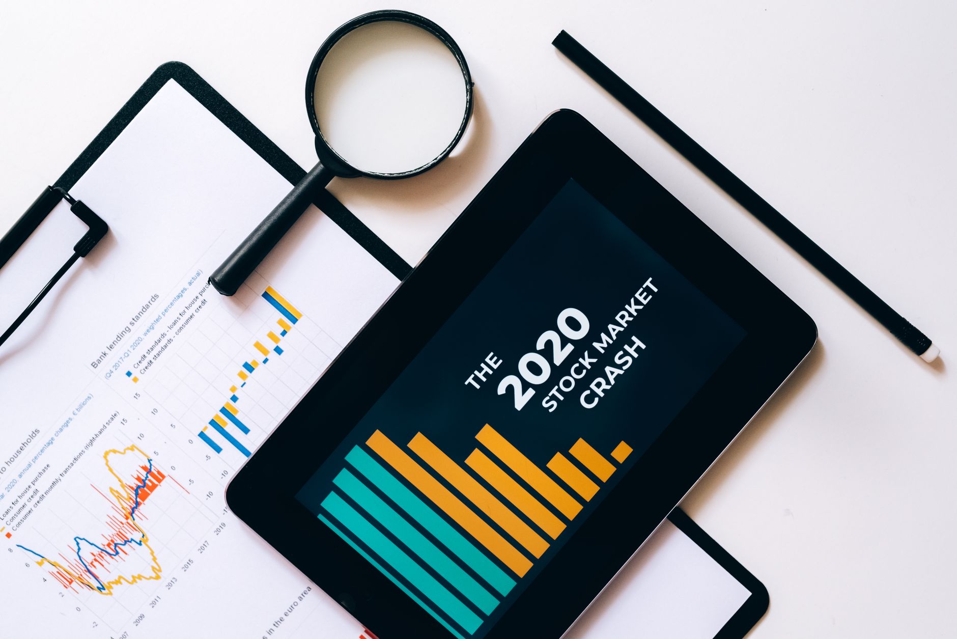 Magnifying glass and tablet analyze 2020 stock market crash data with charts on clipboard.