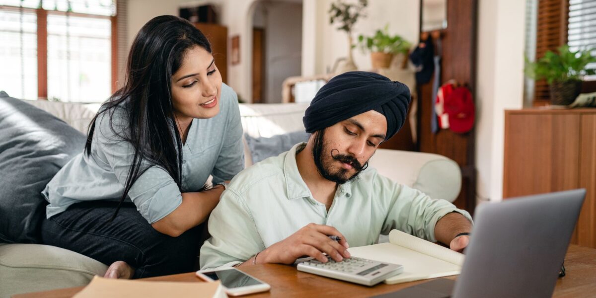 Smiling focused ethnic woman sitting on sofa at home watching working ethnic husband calculating sitting at coffee table among calculator and laptop and smartphone and books and notebook in living room