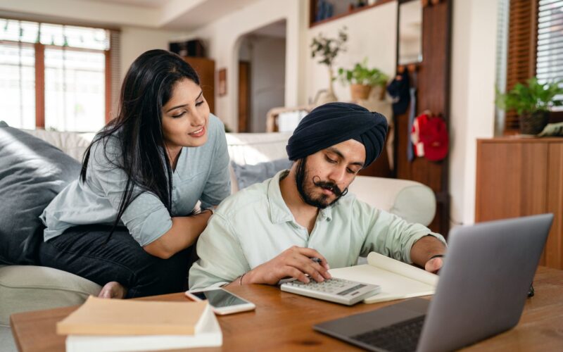 Smiling focused ethnic woman sitting on sofa at home watching working ethnic husband calculating sitting at coffee table among calculator and laptop and smartphone and books and notebook in living room