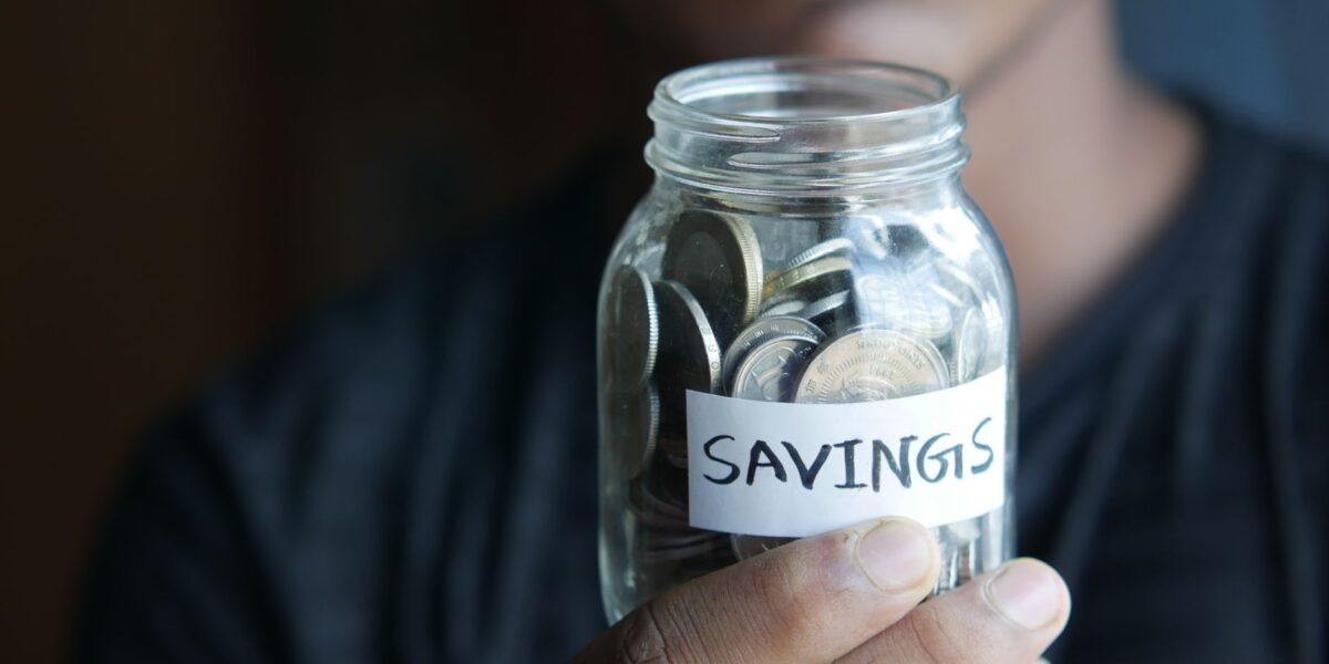 A close-up image of a person's hand holding a jar full of coins labeled 'Savings'.