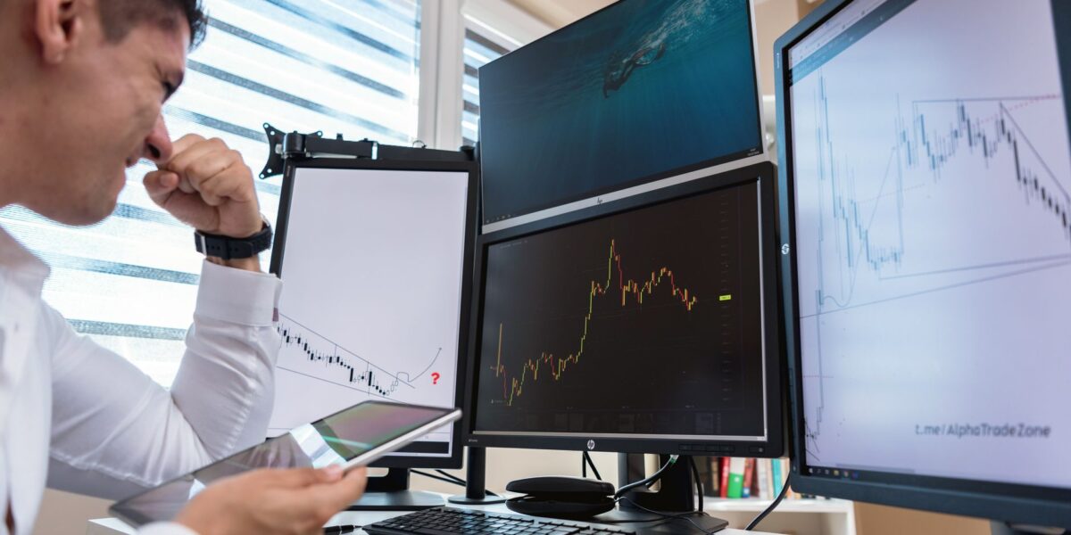 Trader in white shirt analyzing stock charts on multiple monitors during daytime in an office setting.