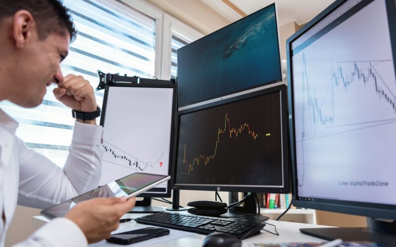 Trader in white shirt analyzing stock charts on multiple monitors during daytime in an office setting.