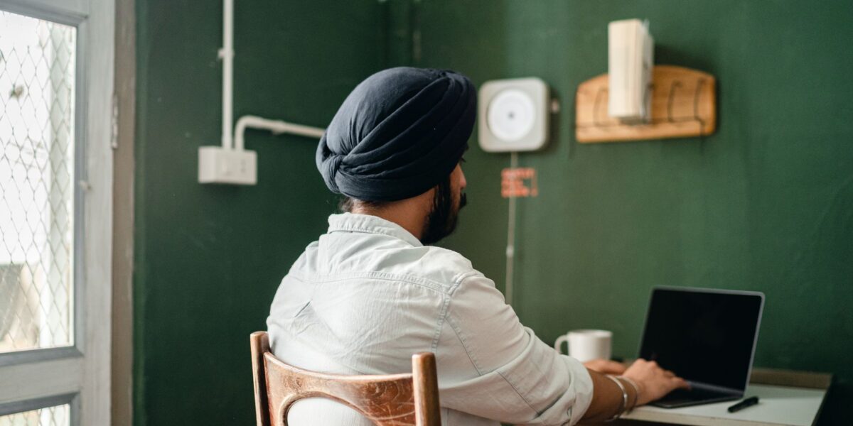 Back view of anonymous bearded Sikh male freelancer in turban and shirt typing on laptop while working on remote project