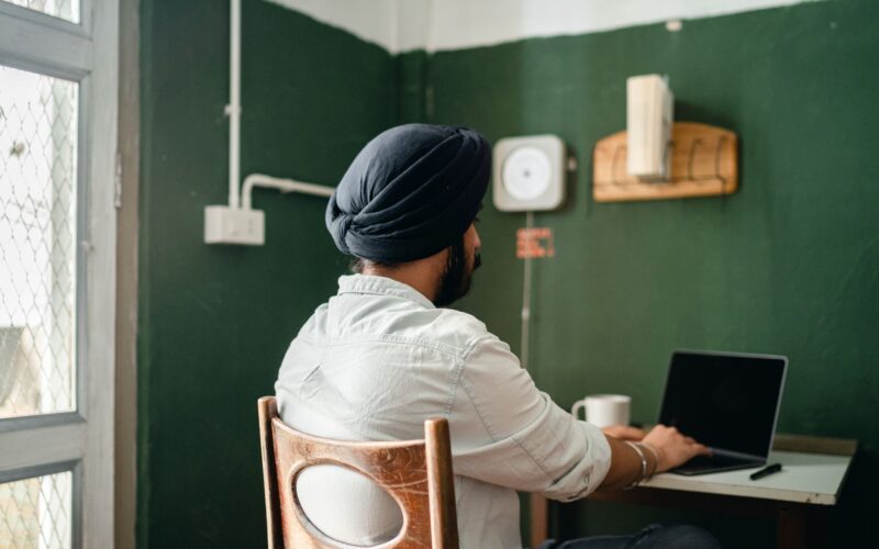 Back view of anonymous bearded Sikh male freelancer in turban and shirt typing on laptop while working on remote project