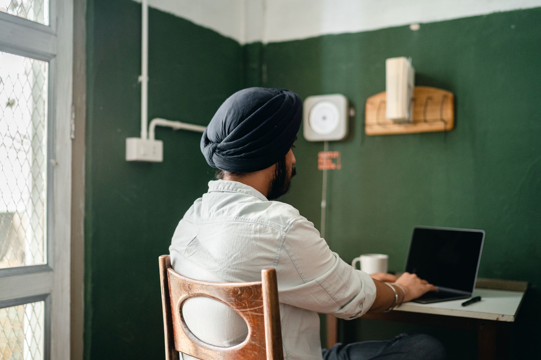Back view of anonymous bearded Sikh male freelancer in turban and shirt typing on laptop while working on remote project