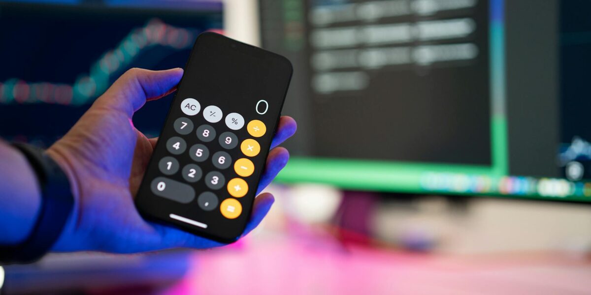 A person uses a smartphone calculator app in a modern office with computer screens displaying charts.