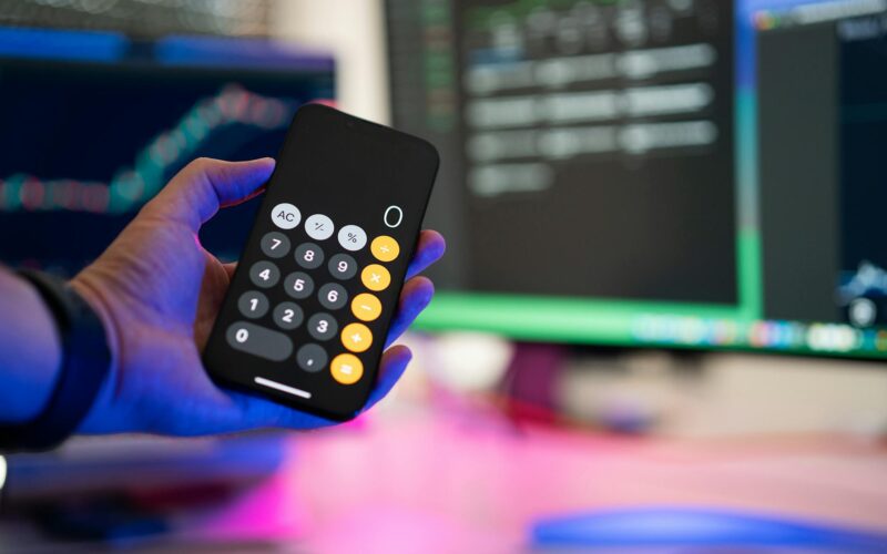 A person uses a smartphone calculator app in a modern office with computer screens displaying charts.