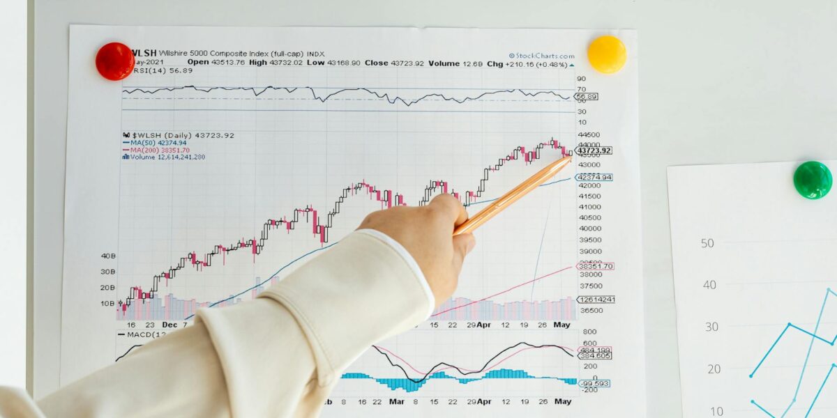 Close-up of a woman's hand pointing to data trends on a stock market chart using a pencil.