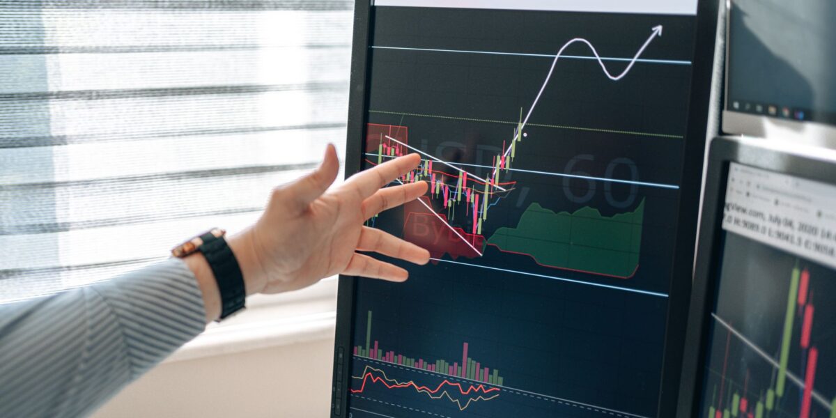 Close-up of a hand pointing at stock market graphs on a monitor in a workspace.