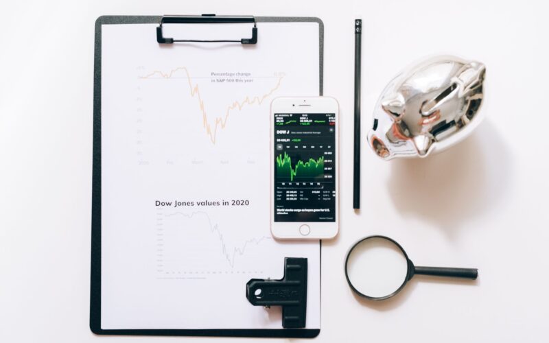 Top view of financial analysis tools including a cellphone, clipboard, magnifying glass, and piggy bank on a white desk.