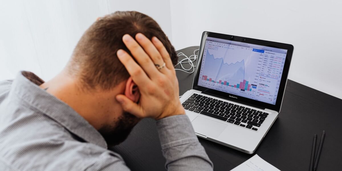 Stressed man at desk looking at declining stock charts on laptop, indicating financial loss.