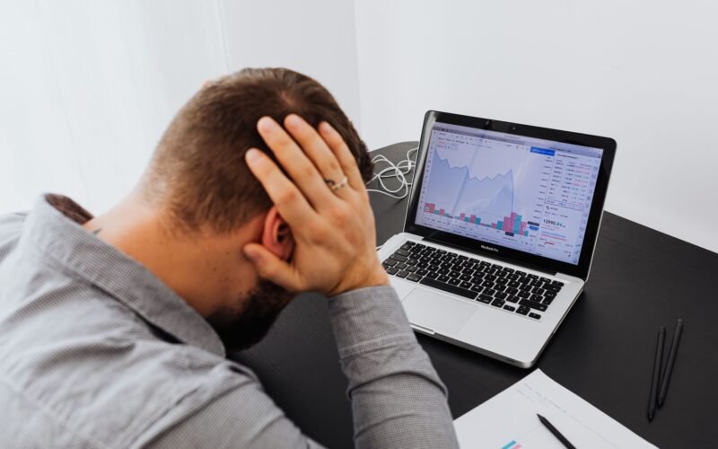 Stressed man at desk looking at declining stock charts on laptop, indicating financial loss.