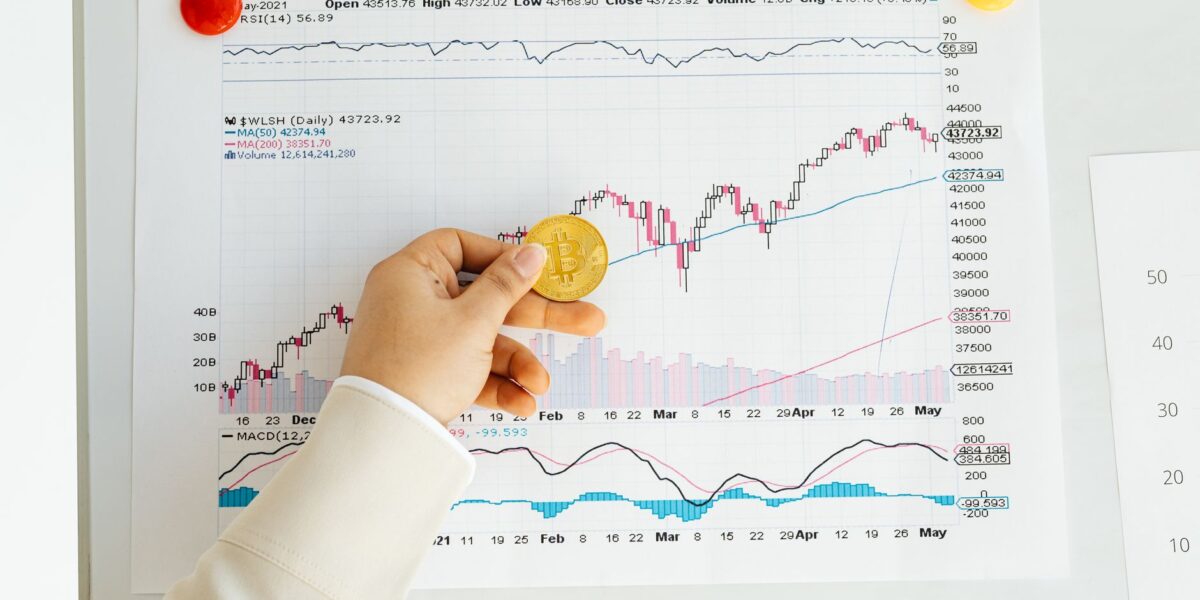 A hand holding a Bitcoin coin in front of a stock market chart, symbolizing analysis and finance.