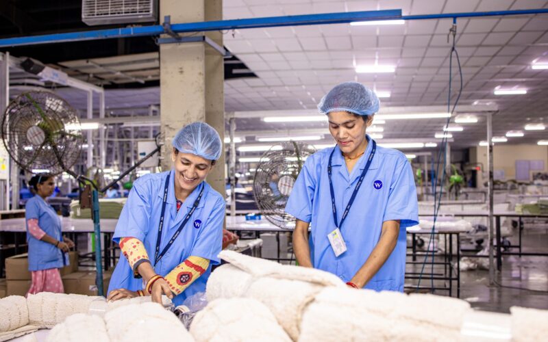 Smiling Indian factory workers in a textile factory wearing blue uniforms and hairnets, working diligently.