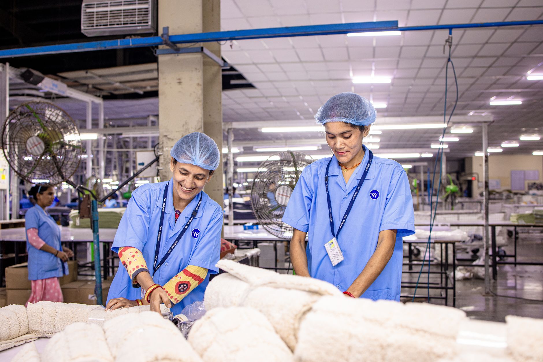 Smiling Indian factory workers in a textile factory wearing blue uniforms and hairnets, working diligently.