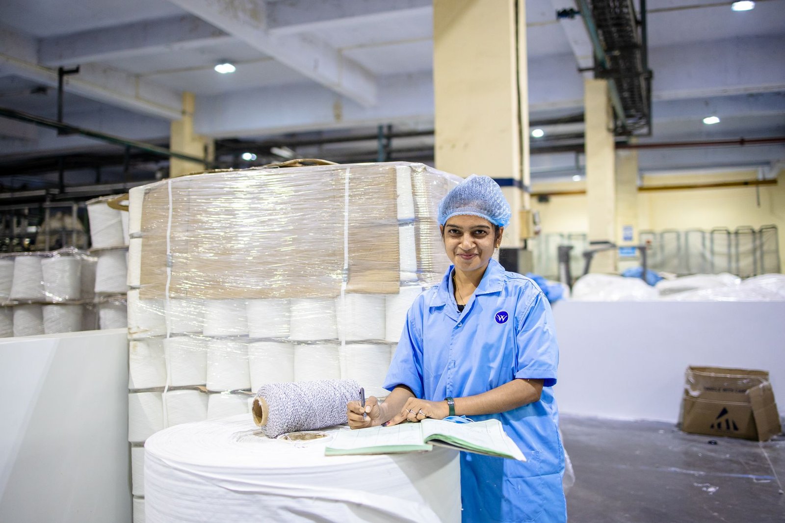 Smiling female textile worker taking notes in a factory with yarn stacks.