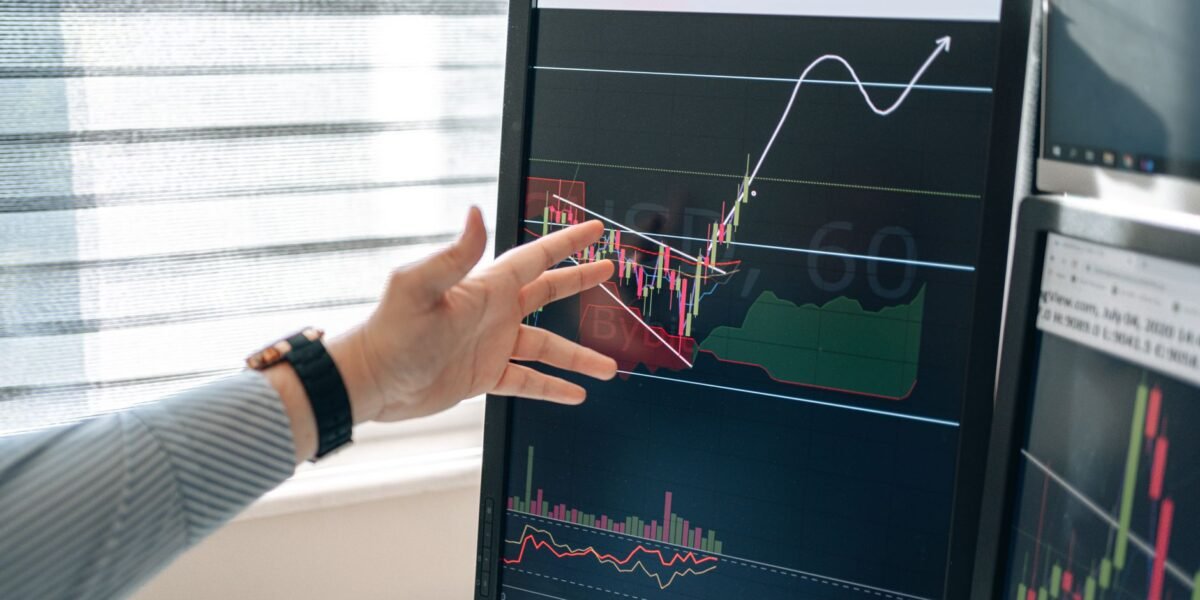 Close-up of a hand pointing at stock market graphs on a monitor in a workspace.