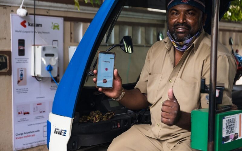 South Asian man using electric vehicle charging station in India, holding smartphone with app.