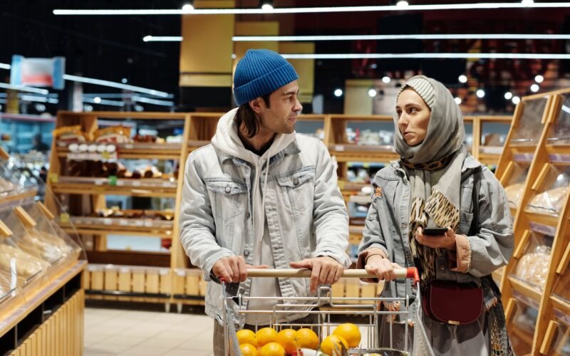 A diverse couple shopping for groceries in a supermarket aisle with a cart full of fruits.