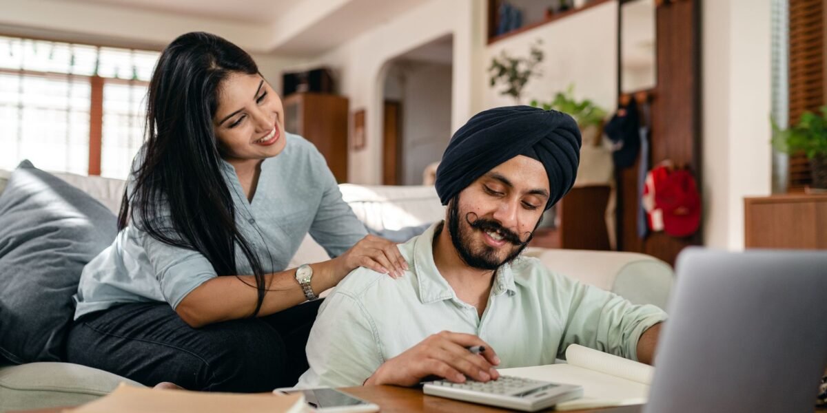 Positive ethnic man sitting at coffee table with laptop and smartphone and books and notebook and calculating while smiling ethnic wife sitting on sofa and supporting and watching behind