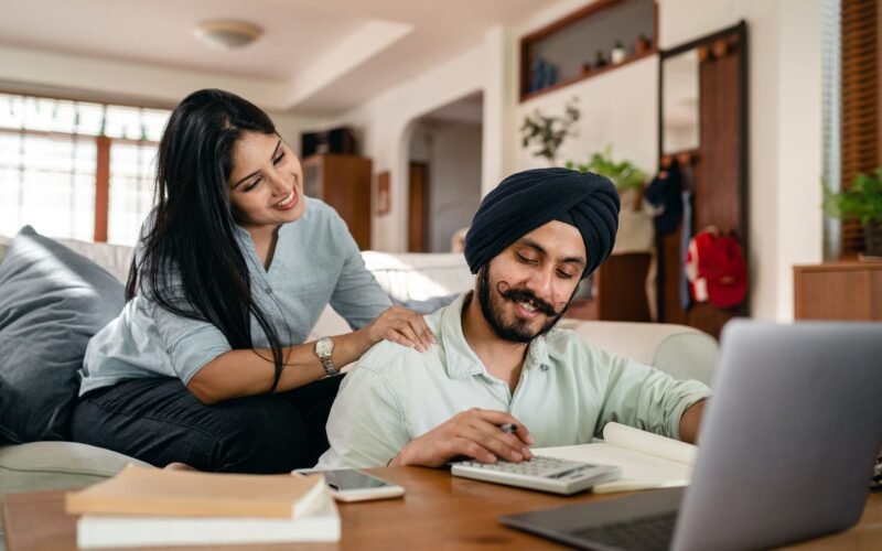 Positive ethnic man sitting at coffee table with laptop and smartphone and books and notebook and calculating while smiling ethnic wife sitting on sofa and supporting and watching behind