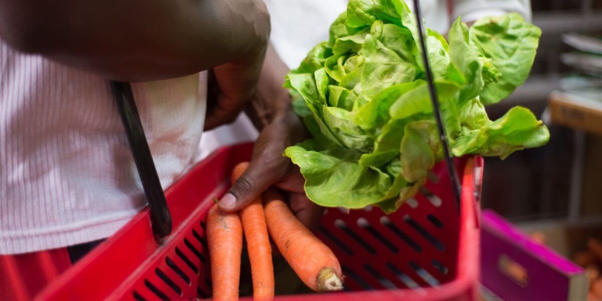 Hands holding red basket with fresh lettuce and carrots in a supermarket setting.