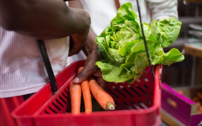 Hands holding red basket with fresh lettuce and carrots in a supermarket setting.
