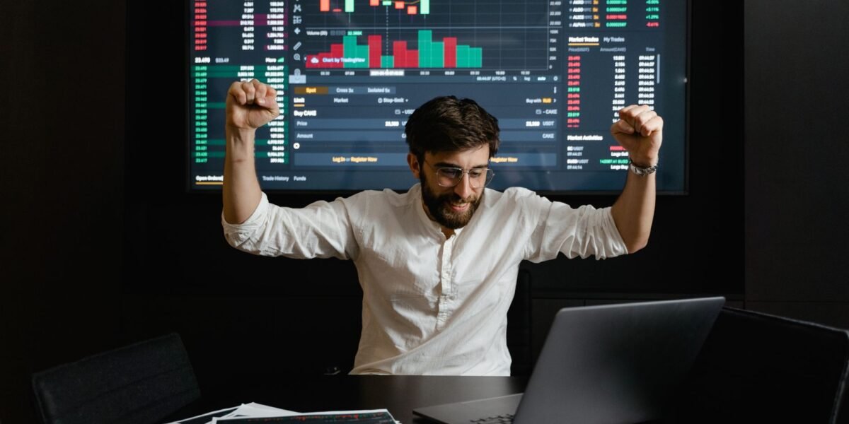 Businessman celebrates stock market success with hands raised in excitement at a trading desk.