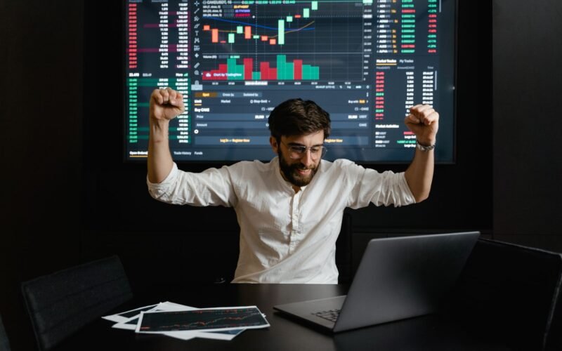 Businessman celebrates stock market success with hands raised in excitement at a trading desk.