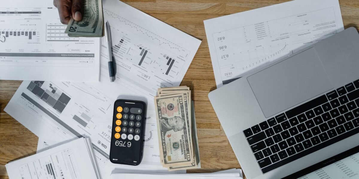 Overhead view of a busy workspace with cash, financial documents, and a laptop.