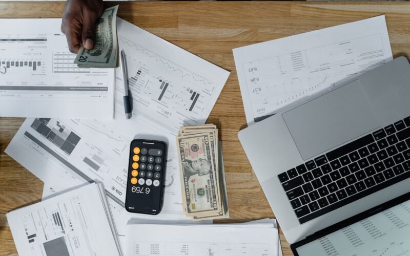 Overhead view of a busy workspace with cash, financial documents, and a laptop.