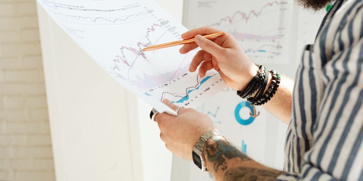 A man analyzing stock market charts with a pen, holding a paper report indoors.