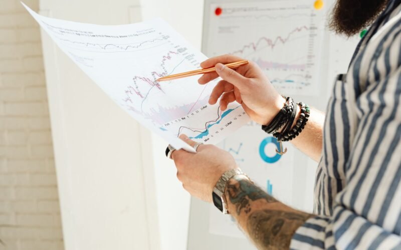 A man analyzing stock market charts with a pen, holding a paper report indoors.