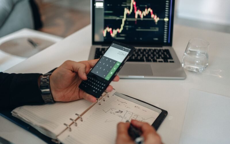 Close-up of a person using a calculator and laptop with stock charts for financial analysis.