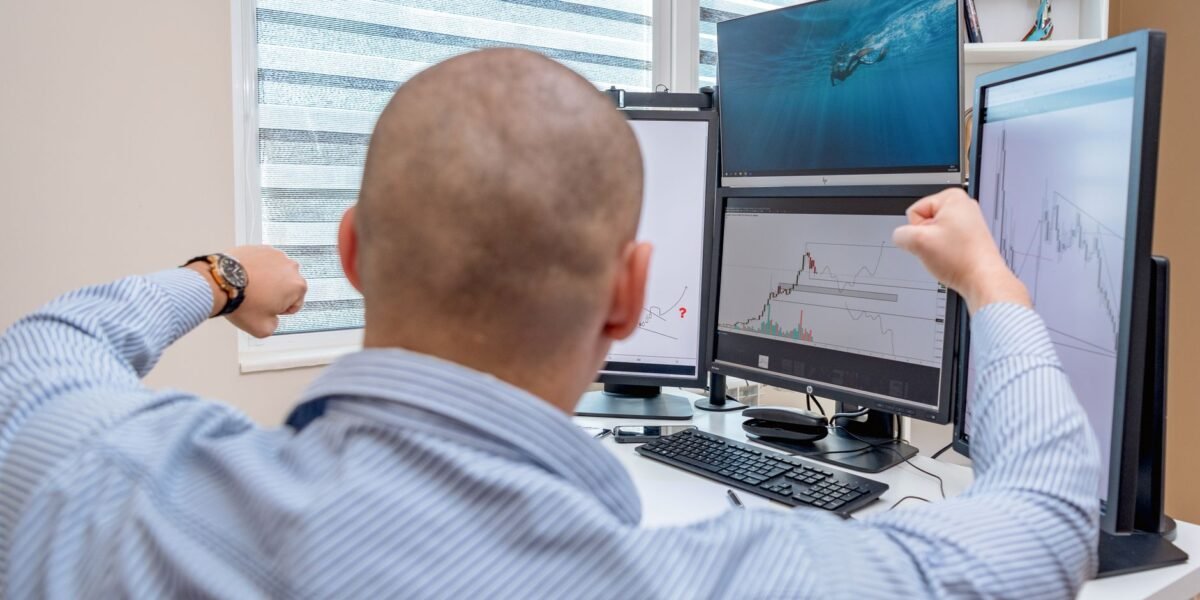 A man celebrates success at a multi-monitor workstation while analyzing stock charts.