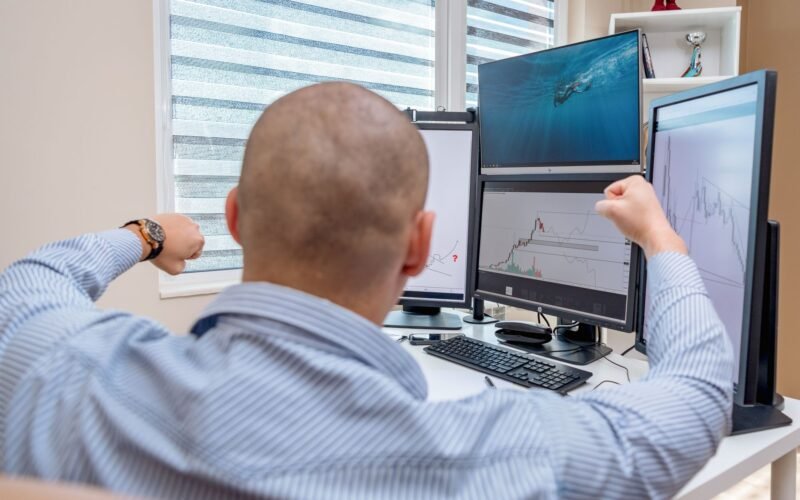 A man celebrates success at a multi-monitor workstation while analyzing stock charts.