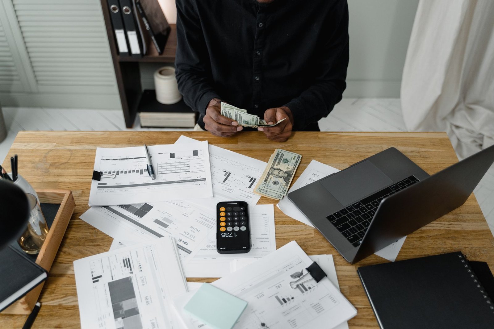 A person counts dollar bills at a desk with a laptop, papers, and calculator.