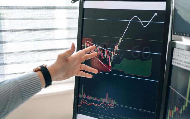 Close-up of a hand pointing at stock market graphs on a monitor in a workspace.