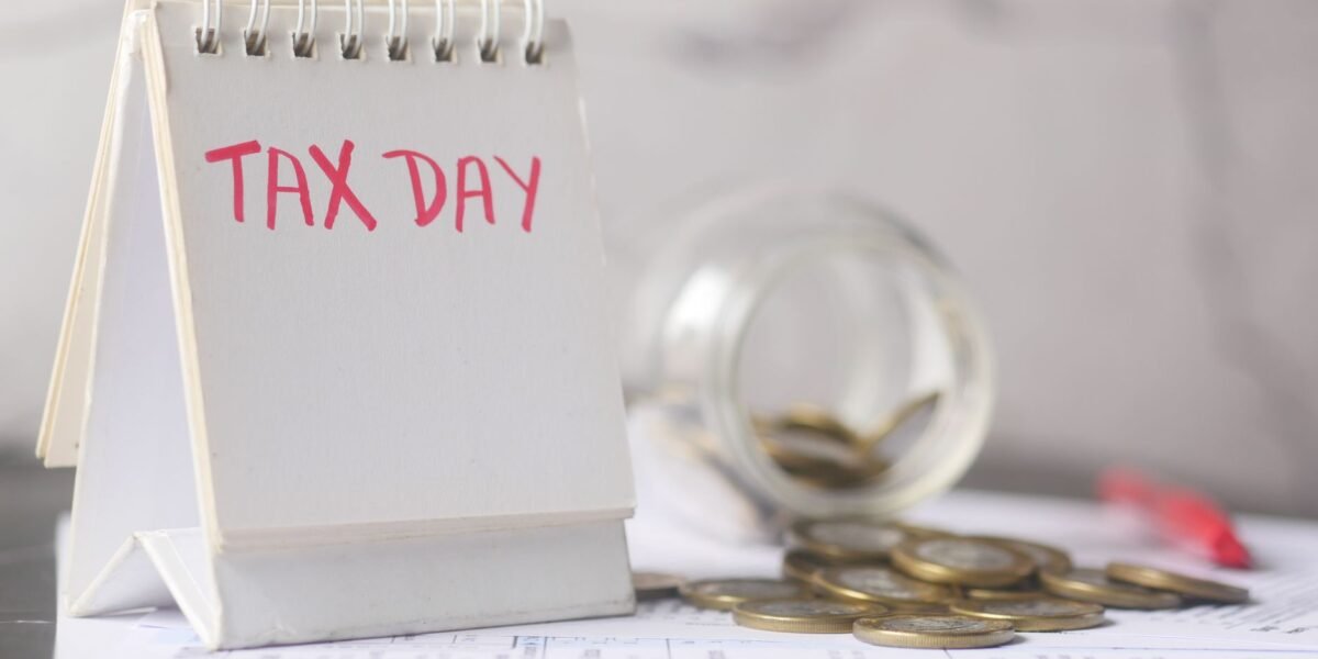 A close-up of a table calendar marked 'Tax Day' with a jar of coins and notes, symbolizing financial planning.