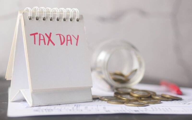 A close-up of a table calendar marked 'Tax Day' with a jar of coins and notes, symbolizing financial planning.
