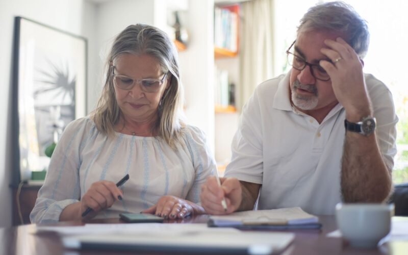 Senior couple calculating expenses at home office desk with documents and notes.