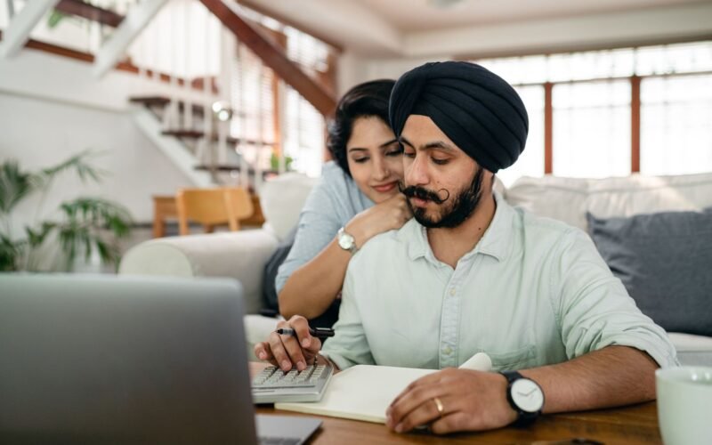 Young smiling woman hugging concentrated husband from behind while counting bills on calculator in modern apartment