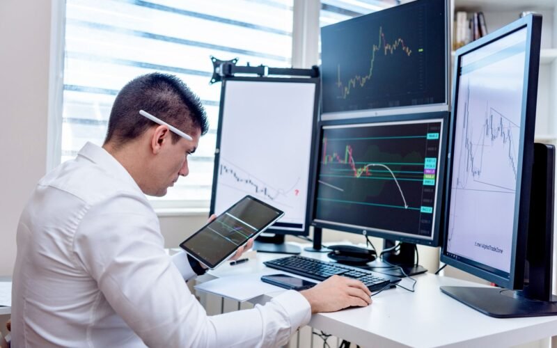 Professional analyzing stock market graphs on multiple monitors at work desk.