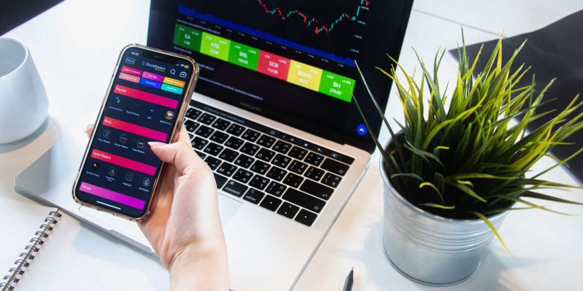 A person using a smartphone app to trade stocks with a laptop displaying market data.