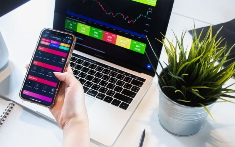 A person using a smartphone app to trade stocks with a laptop displaying market data.