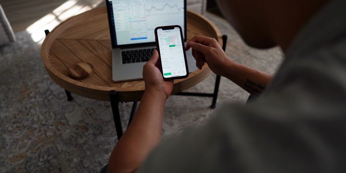 Man trading stocks online using smartphone and laptop. Indoor setting, focus on technology and finance.