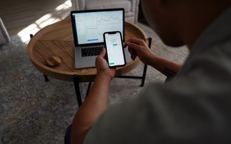 Man trading stocks online using smartphone and laptop. Indoor setting, focus on technology and finance.