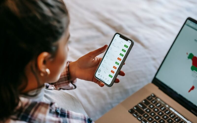From above side view of crop ethnic female broker using financial exchange app on cellphone against netbook at home