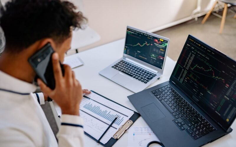 Man analyzing stock market charts on laptops while talking on a cellphone.